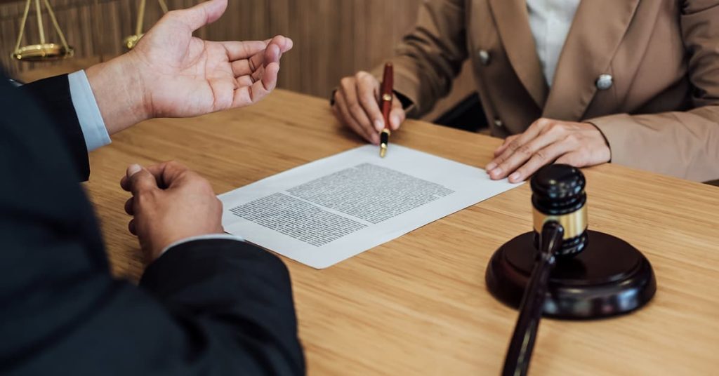 Close-up of two professionals at a wooden desk, one pointing at a densely written legal document while the other listens. A gavel and scales of justice are visible, indicating a legal or courtroom environment. The scene suggests a consultation or explanation of legal terms before signing.