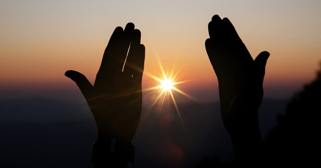 Silhouette of two hands raised in a prayerful gesture against a glowing sunrise. The sun shines brightly between the palms, casting rays of light across a serene gradient sky transitioning from deep orange to soft blue. The peaceful scene evokes a sense of spirituality and reflection