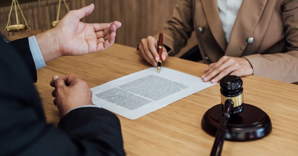 Two professionals sit across from each other at a wooden desk discussing a legal document. One person holds a pen, ready to sign, while the other gestures in explanation. A judge’s gavel and scales of justice are visible on the desk, indicating a legal or courtroom setting