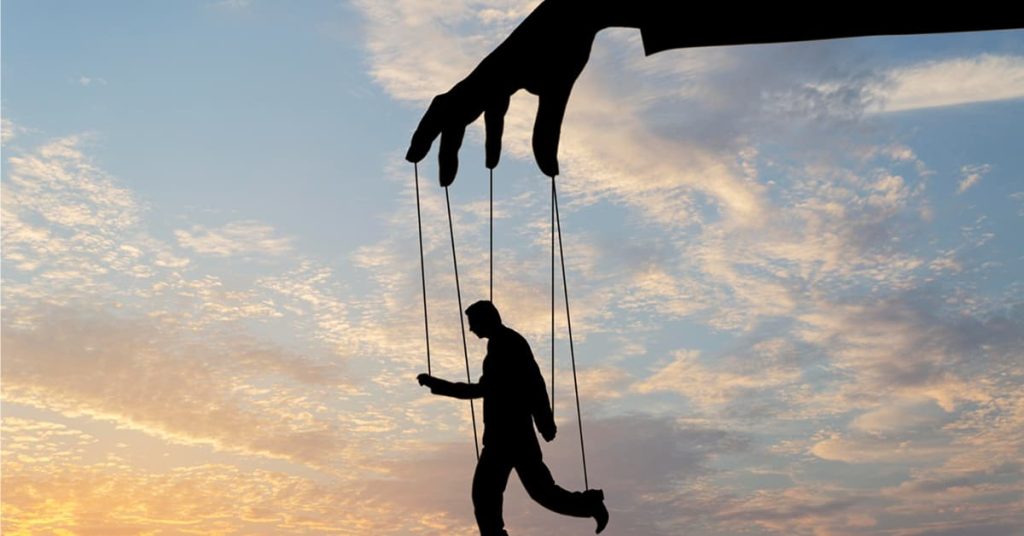 A conceptual image showing the silhouette of a large hand controlling a puppet figure with strings against a serene sky at sunset. The puppet appears to walk, symbolizing control, manipulation, or lack of free will. The soft, colorful clouds contrast the dark figures, emphasizing the metaphor.
