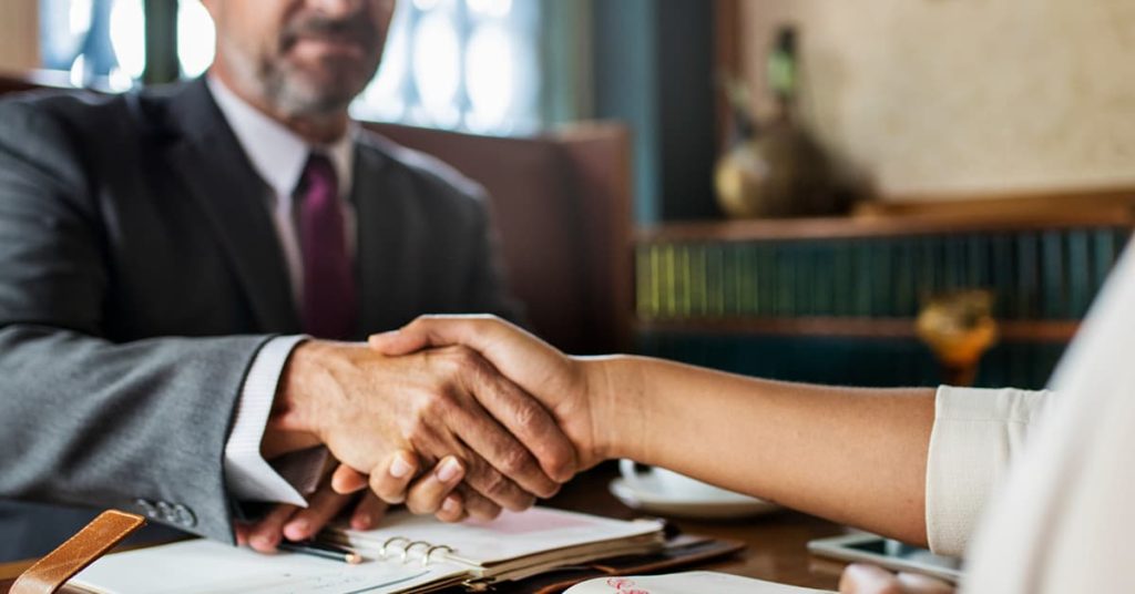 Close-up of a handshake between a man in a suit and a woman in business attire, symbolizing a formal agreement or successful deal. The setting appears to be a professional or business meeting, with a notebook and documents on the table, and a blurred background of office decor.