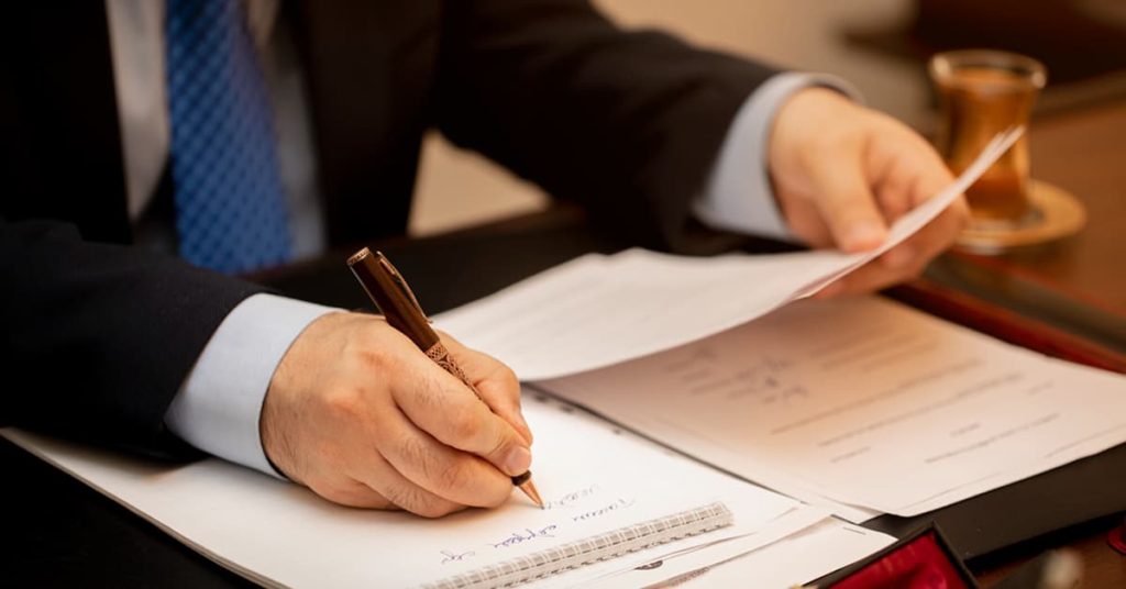 A close-up view of a man in a suit and tie signing documents at a desk. He holds a pen in one hand and a sheet of paper in the other, with several papers and a notepad spread out before him. A gavel and a tea glass are visible in the background, suggesting a formal or legal setting.