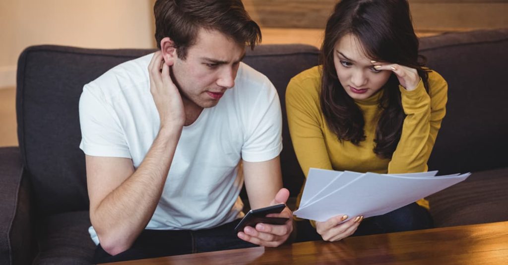 A young couple sits on a couch looking stressed and concerned as they review paperwork. The man holds a smartphone while the woman looks over several documents, her hand to her forehead in frustration. The image conveys financial stress, debt, or tax-related anxiety.