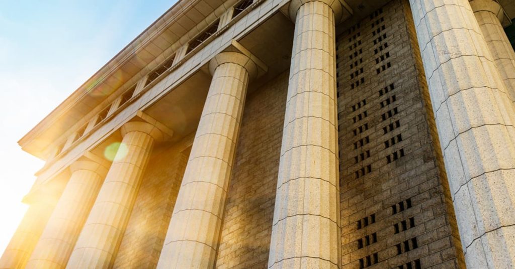 A dramatic upward view of a grand courthouse with tall, classical stone columns and a stone brick facade. The morning or afternoon sun casts a golden glow and lens flare across the structure, emphasizing its architectural strength and formality.